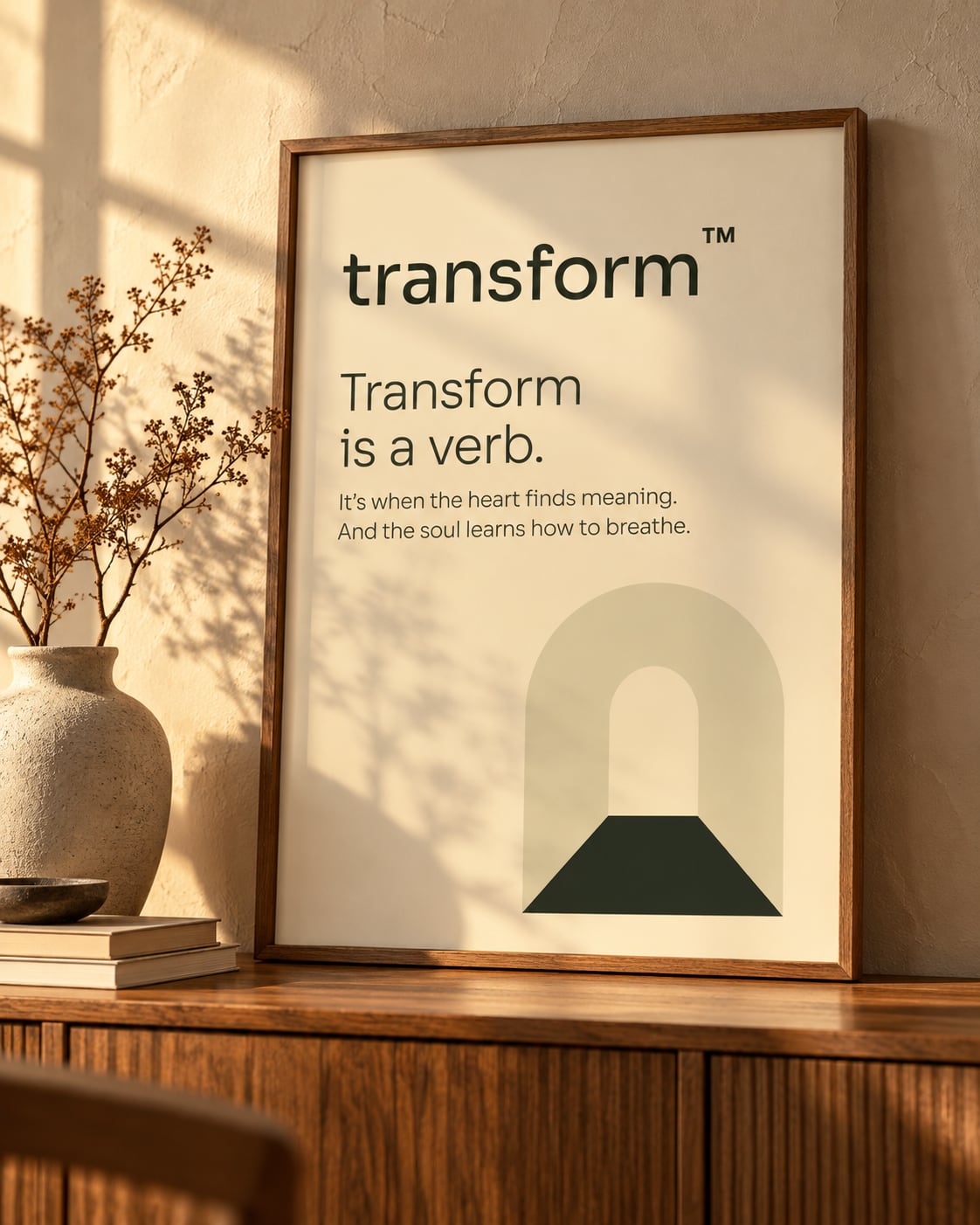 Pôster emoldurado "Transform is a verb." com o símbolo, apoiado num aparador com vaso de flores secas.